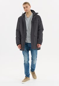 Mörkgrå isolerad jacka med huva, dragkedja fram och flera fickor. Buren över en ljusgrå t-shirt, tillsammans med blå jeans och beige sneakers.