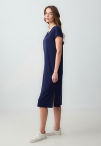 Abito in cotone blu navy con scollo a V, maniche corte e fessure laterali, abbinato a sneaker bianche su uno sfondo neutro.