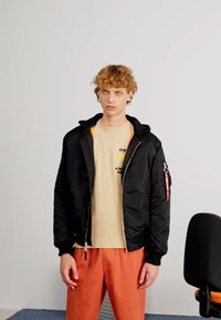 Zwarte bomberjack met een capuchon en oranje voering, gedragen over een beige shirt, gecombineerd met oranje broek. Voorzien van een ritszakdetail.
