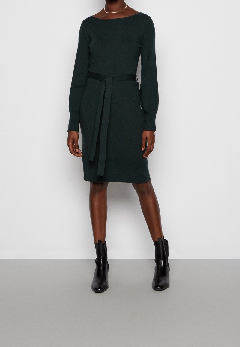 Femme portant une robe ceinturée vert foncé à manches longues, longueur genou, et des bottines noires, debout devant un fond gris clair uni.