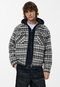 Giacca a quadri bianca e nera con due tasche sul petto, bottoni a pressione e uno strato interno con cappuccio foderato, abbinata a pantaloni di denim scuro.