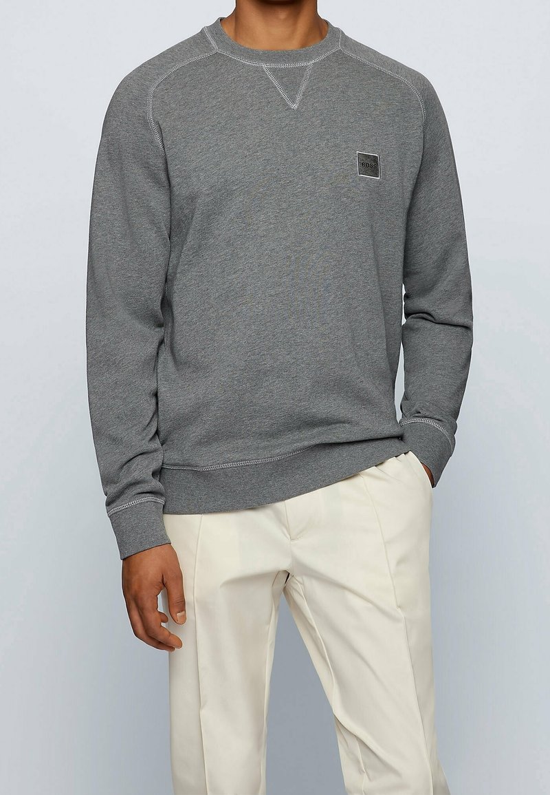 Sweatshirt grigio con scollatura a girocollo, polsini e orlo a costine, cuciture a contrasto e una piccola etichetta con logo sul petto. Abbinato a pantaloni beige.