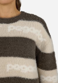 Απαλή ζακέτα με εναλλασσόμενες σκούρες γκρίζες και κρεμ ρίγες με μαλακή υφή. Περιλαμβάνει ένα διακριτικό σχέδιο λογότυπου σε ανοιχτό ύφασμα.