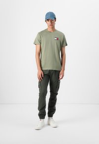 Olivegrünes T-Shirt mit kleinem Logo auf der Brust, kombiniert mit taillierten olivfarbenen Cargo-Hosen und weißen Sneakern. Eine Denim-Cap rundet den Look ab.