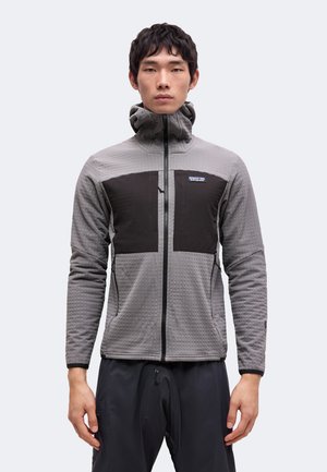 Jeune homme portant une veste grise zippée avec des panneaux noirs sur la poitrine et un col montant, debout devant un fond clair uni.