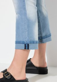 Jeans de corte cropped en azul claro con puños enrollados y una raya azul oscura. Llevados con sandalias negras tipo slide sobre una superficie texturizada.