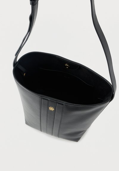 Μαύρη δερμάτινη τσάντα ώμου με μία λουρίδα, τρεις κάθετες ραφές και ένα μικρό χρυσό έμβλημα στο μπροστινό μέρος.