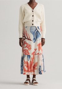 Cardigan a coste beige chiaro con bottoni, abbinato a una gonna fluida color crema con motivi ispirati al mare in blu e corallo, indossato con sandali bianchi.