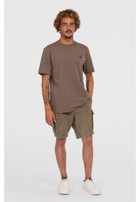 Brun bomulds t-shirt med rund hals og et logo patch på venstre bryst, parret med olivengrønne cargo shorts og hvide sneakers.