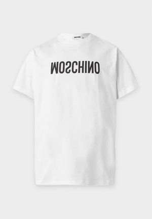 Jonge man staat met handen in de zakken van zwarte broek, draagt een wit T-shirt met omgekeerde tekst en zwarte veterlaarzen.