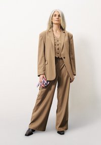 Brauner maßgeschneiderter Anzug mit weit geschnittenen Hosen und einer figurbetonten Weste. Enthält einen langen Blazer und ein farbenfrohes, gemustertes Taschentuch.