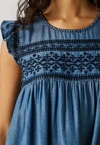 Modra kavbojka obleka z vezenimi geometrijskimi vzorci na prsih in nabrane kratke rokavice na zgornjem delu ženske postave.