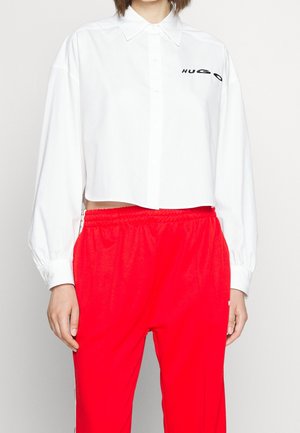 Zuschnitt weißes Hemd mit langen Ärmeln und "HUGO"-Logo, kombiniert mit hoch taillierten, elastischen Hosen in leuchtendem Rot.