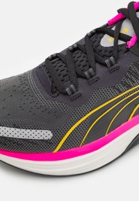 Scarpa da corsa nera con una parte superiore testurizzata, accenti rosa e gialli, lacci neri e una suola intermedia bianca con un design ondulato.