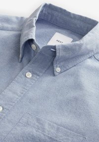 Camisa de color azul claro con cuello, hecha de tejido texturizado, que presenta un bolsillo en el pecho, botones blancos y una pequeña etiqueta de la marca en el interior del cuello.