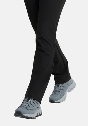 Legs wearing black pants and light gray hiking shoes with thick, rugged soles stepping forward on a plain background.