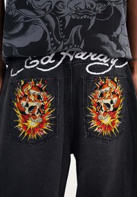 Black denim pants featuring embroidered skull designs with flame accents on back pockets, and a white embroidered brand name at the waistband.