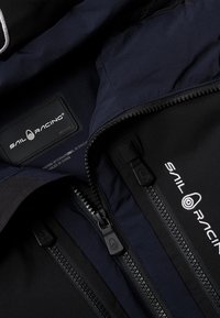 Marinblå jacka med kontrasterande svarta detaljer, med flera dragkedjor, logotypmärke och slätt, strukturerat tyg. Inkluderar en huva.