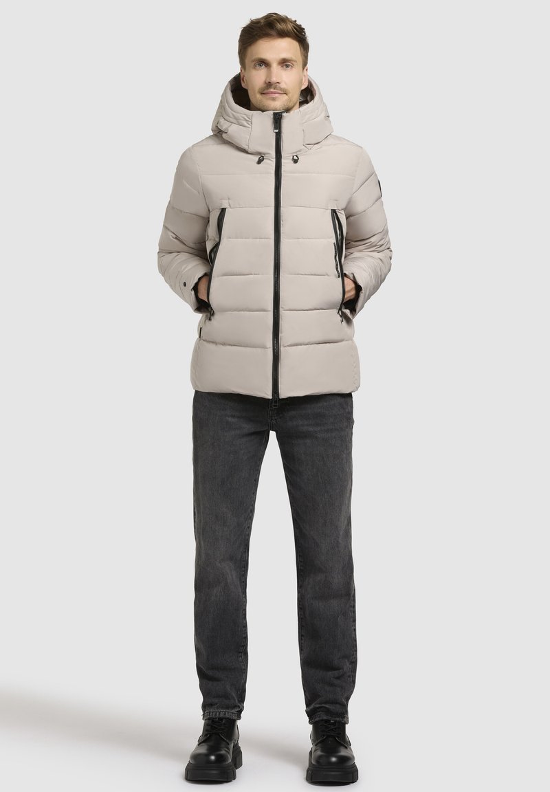 Bežna pufera jakna s kapuco, ki ima črne zadrge in kviltano obliko. V kombinaciji s temnimi kavbojkami in črnimi čevlji.