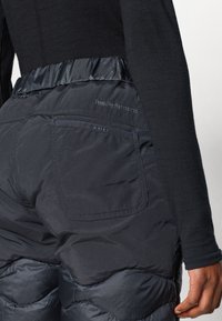 Calças pretas acolchoadas com cintura ajustada e bolsos laterais; textura leve e suave; apresenta detalhe do logótipo Peak Performance.