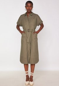 Donna nera in piedi con le mani nelle tasche, che indossa un vestito midi verde oliva con cintura e scarpe espadrillas beige con cinturini alla caviglia, contro un muro bianco.