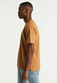 Brun bomulds t-shirt med korte ærmer, rund halsudskæring og sideslidser, båret med lyseblå denimjeans, der viser et enkelt design.