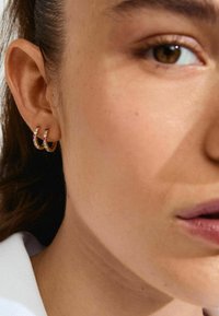 Pendientes de aro de oro con piedras preciosas coloridas; el primer plano muestra detalles intrincados y textura sobre un tono de piel neutro.