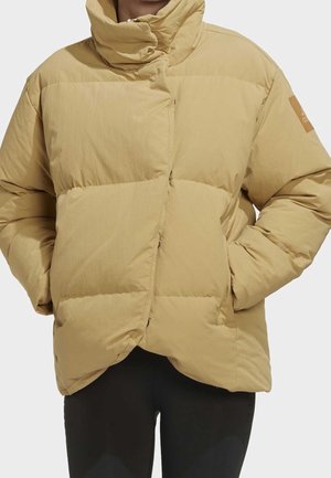 Veste d'hiver matelassée beige avec col montant, manches longues et mains dans les poches latérales, portée avec un pantalon noir.