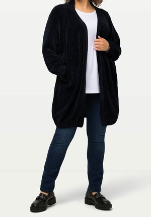 Frau trägt schwarzen, strukturierten, offenen Cardigan, weißes Hemd, dunkelblaue Jeans und schwarze klobige Loafers, steht vor einfachem Hintergrund.