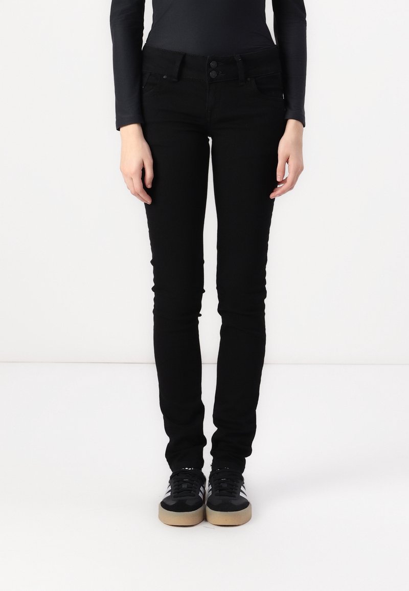 Person som bär svarta skinny jeans och svarta sneakers med vita ränder, stående mot en vit bakgrund.