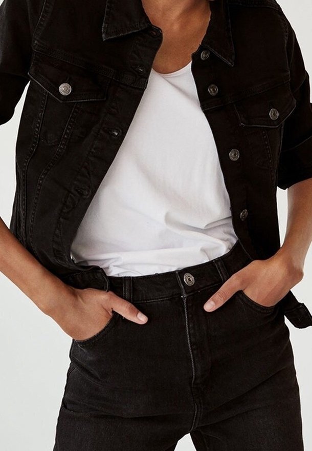 Chaqueta de mezclilla negra con botones plateados, combinada con una camiseta blanca y jeans negros de cintura alta. Diseño casual y ajustado con bolsillos.