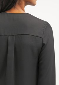 Blusa nera con una texture liscia, caratterizzata da una semplice scollatura rotonda, dettaglio a pieghe e maniche lunghe. Il tessuto appare leggero e morbido.