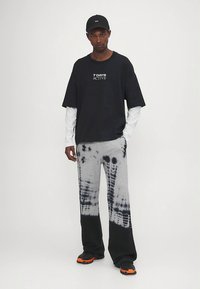 Must graafiline t-särk tekstiga "7 DAYS ACTIVE", kantud valge pika varrukaga särgi peal. Hallid tie-dye dressipüksid ja mustad tossud oranžide detailidega.