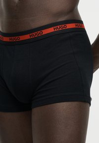 Ανδρικός κορμός που φορά μαύρα boxer briefs με κόκκινη μέση που φέρει το επαναλαμβανόμενο όνομα της μάρκας "HUGO" με μαύρα γράμματα.