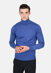 Maglione a collo alto blu in morbido tessuto a maglia. Presenta maniche lunghe e polsini a coste, indossato con pantaloni neri su misura.
