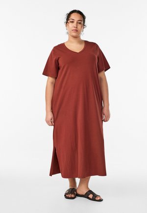 Naine seisab, kandes roostepunast lühikeste varrukatega pahkluud ulatuvat kleiti külje lõhikute ja mustade libisevate sandaalidega valgel taustal.