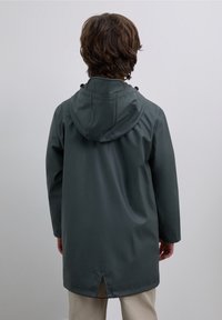 Mörkgrön vattentät jacka med huva, långa ärmar och en ryggventil. Slät, blank yta med dragsko vid kragen.