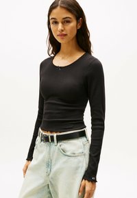 Top a maniche lunghe nero a coste con scollo rotondo, orlo ondulato e un piccolo fiocco viola decorativo sul colletto. Indossato con jeans azzurri chiari.