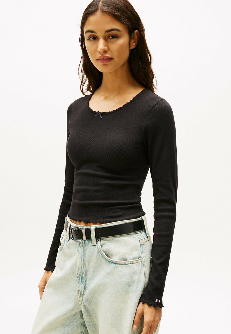 Top a maniche lunghe nero a coste con scollo rotondo, orlo ondulato e un piccolo fiocco viola decorativo sul colletto. Indossato con jeans azzurri chiari.