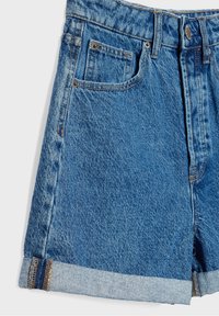Pantalones cortos de mezclilla en un lavado azul medio con un acabado ligeramente descolorido. Presentan un dobladillo acampanado, bolsillos frontales y acentos de hardware metálico.