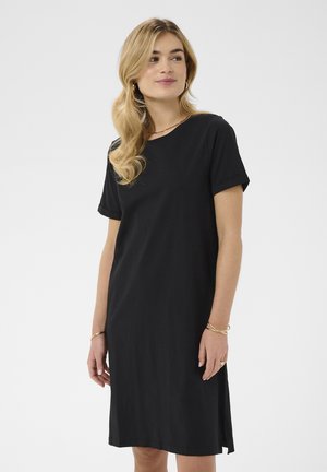 Žena s vlnitými blond vlasy, oblečená v černých šatech s krátkým rukávem, zlatými kruhovými náušnicemi a zlatými náramky, dívající se stranou s jemným úsměvem.