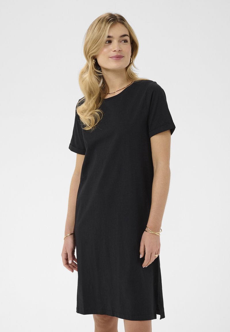 Kobieta o falistych blond włosach, ubrana w czarną sukienkę z krótkim rękawem, złote kolczyki koła i złote bransoletki, spoglądająca na bok z lekkim uśmiechem.