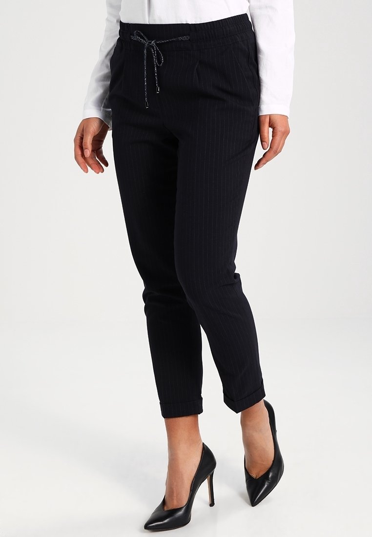 Femme portant un pantalon noir slim à fines rayures avec ourlets retroussés, des talons hauts noirs et un haut à manches longues blanc sur fond blanc.