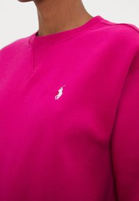 Cerise sweatshirt i mjukt material med rund halsringning och en liten vit broderad logotyp på bröstet.