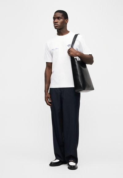 Νέος άνδρας που στέκεται, φοράει λευκό μπλουζάκι με σχέδιο, μπλε παντελόνι με ρίγες και φαρδιά μπατζάκια, μαύρα και άσπρα μοκασίνια, κρατώντας μαύρη δερμάτινη τσάντα ώμου.