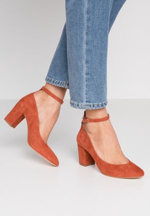 Roodbruine suède blokhakken met enkelbandjes, gedragen met lichtblauwe denim jeans tegen een effen grijze achtergrond.