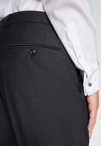 Donkergrijze op maat gemaakte broek met een gladde textuur, voorzien van een achterzak met een glanzende knop. Een uiteinde van een wit overhemd is zichtbaar.