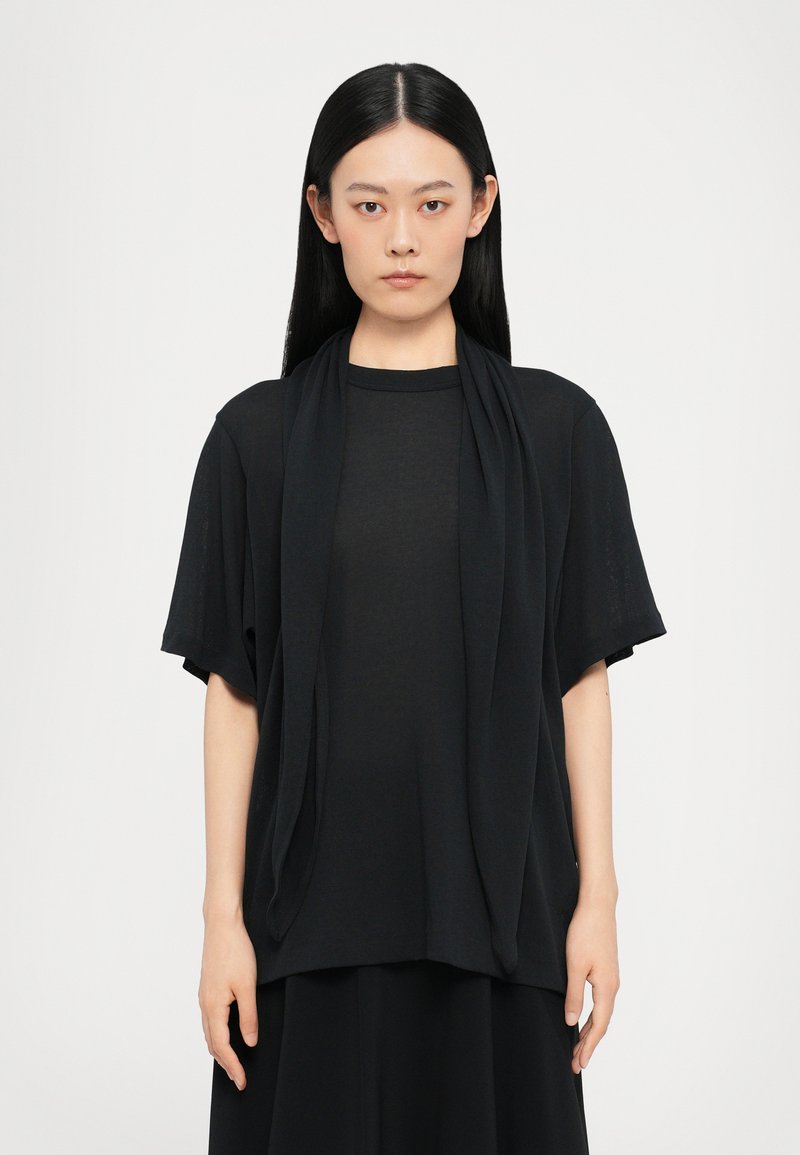 Top nero a maniche corte con design drappeggiato, dotato di un collo rotondo e una consistenza morbida e fluida. Indossato sopra una lunga gonna nera coordinata.