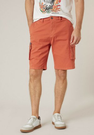 Homme portant un short cargo orange avec poches avant et des baskets blanches, visible de la taille aux pieds sur un fond uni.