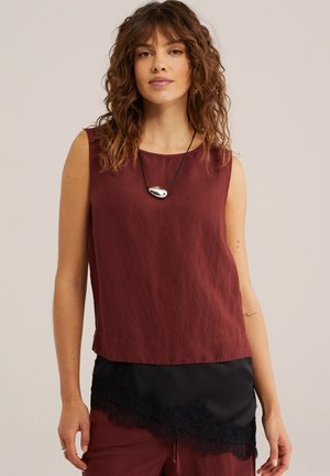 Femme aux cheveux bouclés portant un haut rouge foncé sans manches et une jupe noire à volants bordée de dentelle, accessoirisée d’un pendentif argenté sur un cordon noir.